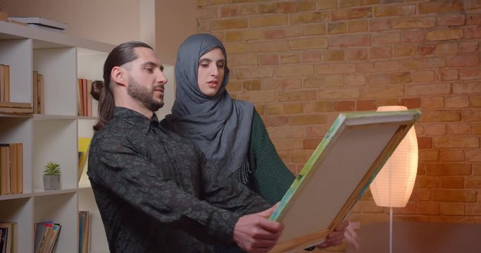 Closeup shoot of young attractive muslim couple settling in a newly bought apartment indoors. Man and female advicing on where to hand a picture