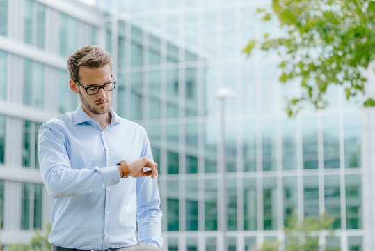 Young businessman standing in front of modern office building, checking the time