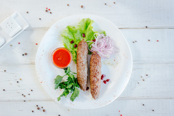 Meat kebabs on white plate with green herbs and red sauce.