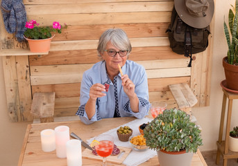 Front view of elderly woman gray hair who enjoys aperitif eating salami and olives. Sitting on a...