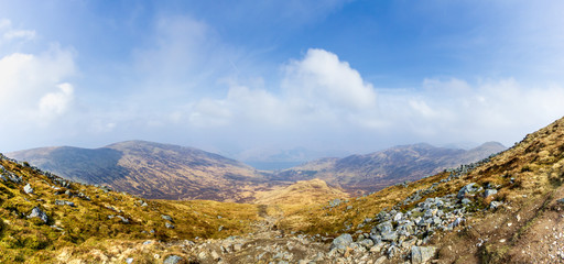 A view of a mountain valley with rocky path,grass and lake in the background under a majestic blue sky and white clouds