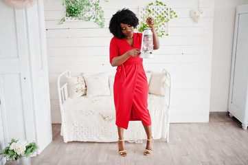 Fashion african american model in red beauty dress, sexy woman posing evening gown in white vintage room with old lantern in hands.