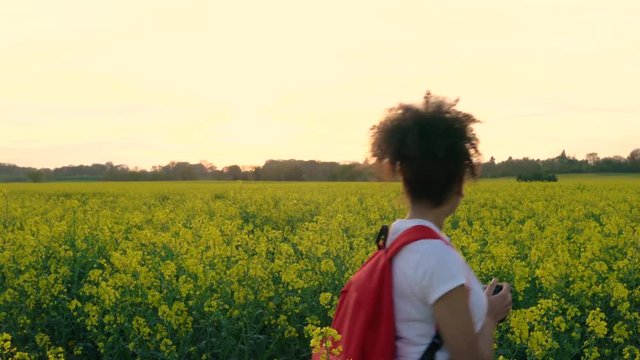 4K video clip of healthy mixed race African American girl teenager female young woman photographer with red backpack and camera taking photographs in field of yellow flowers