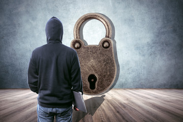 back view of a hacker in the hood with a laptop in hand, in an empty room in front of a huge lock,...