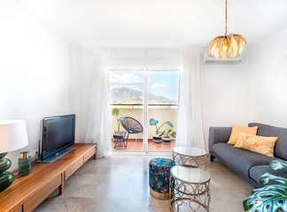 Bright living room with sofa, wooden table, side tables, tv,plants and big window to a terrace with mountain view. Summer holidays vacation.Spacious modern interior
