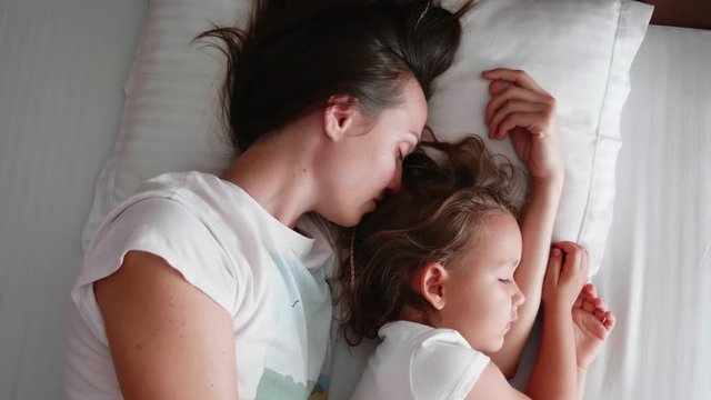 Family portrait of young mom is sleeping with her cute little daughter together on white linen in bed.