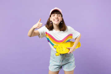 Portrait of smiling teen girl in vivid clothes with yellow skateboard, showing thumb up isolated on violet pastel background in studio. People sincere emotions, lifestyle concept. Mock up copy space.