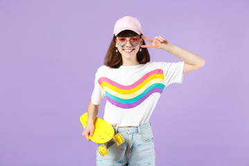 Pretty teen girl in vivid clothes heart eyeglasses hold yellow skateboard showing victory sign isolated on violet pastel wall background. People sincere emotions lifestyle concept. Mock up copy space.