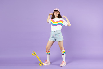 Fototapeta na wymiar Happy teen girl in vivid clothes standing with skateboard keeping eyes closed doing winner gesture isolated on violet pastel background. People sincere emotions lifestyle concept. Mock up copy space.