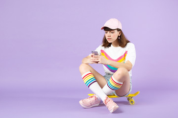 Teen girl in vivid clothes sitting on yellow skateboard, using mobile phone, typing sms message isolated on violet pastel background. People sincere emotions, lifestyle concept. Mock up copy space.