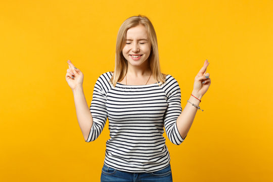 Smiling young woman waiting for special moment, keeping fingers crossed, eyes closed, making wish isolated on yellow orange background. People sincere emotions, lifestyle concept. Mock up copy space.