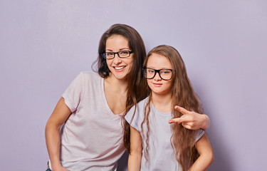 Happy fun young casual mother showing the horns sign two fingers and hugging her cute kid girl on violet wall background. Family in fashion eye glasses.