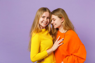 Two smiling young blonde twins sisters girls in vivid colorful clothes hugging looking camera aside isolated on pastel violet blue wall background. People family lifestyle concept. Mock up copy space.