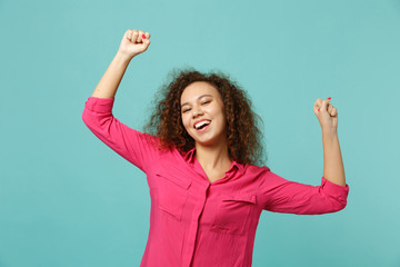 Portrait of joyful african girl in pink casual clothes clenching fists like winner isolated on blue turquoise wall background in studio. People sincere emotions, lifestyle concept. Mock up copy space.