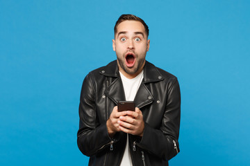 Handsome fun overjoyed excited young unshaven man in black leather jacket white t-shirt using mobile phone isolated on blue wall background studio portrait. People lifestyle concept Mock up copy space