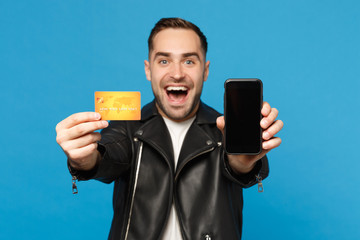 Young man in black jacket white t-shirt hold mobile phone empty screen for promotional content credit card isolated on blue wall background studio portrait. People lifestyle concept Mock up copy space