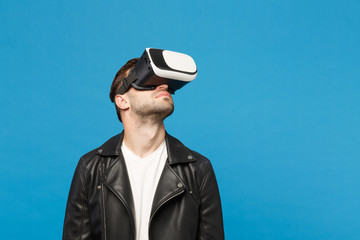 Stylish young unshaven man in black jacket white t-shirt looking in headset, virtual reality vr isolated on blue wall background studio portrait. People emotions lifestyle concept. Mock up copy space.