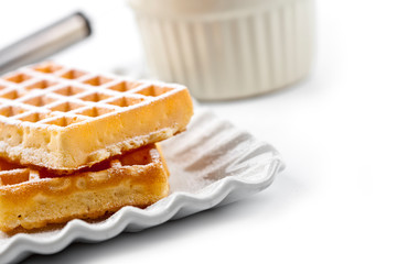 Belgium waffers with sugar powder on ceramic plate and strainer on white table.