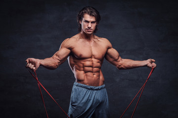 Fototapeta premium Attractive shirtless man is looking to the camera while doing exercises with rubber. There is dark background.