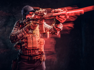 Fototapeta na wymiar Special forces soldier wearing a checkered shirt and protective equipment holding an assault rifle and aim at the enemy. Red light effect in motion. Studio photo against a dark textured wall