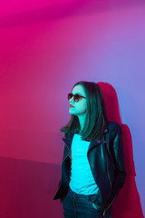 Fashioned girl in leather jacket in dark glasses in pink anf blue  neon lights background. 