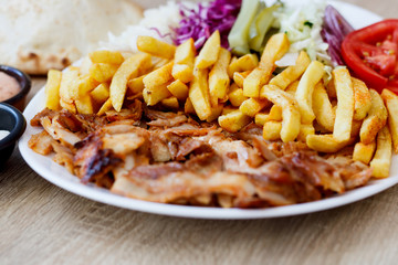 Chiken Doner Kebab on the plate with french fries, tomatoes, onion, pickles and salad on a wooden background. Grilled chicken meat with vegetables.