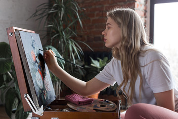 Young pretty girl with brush and palette sitting near easel drawing picture. Art, creativity, hobby, drawing process.