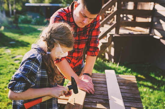 Father Teaching Kid Daughter To Work With Wood Outdoor. Dad Explaining How To Use Hammer And Build Wooden Details. Country House On Background