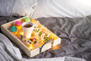 Morning breakfast in bed. Wooden tray with coffee or tea and cupcakes. Gray bedspread in the interior of a hotel room. Copy space