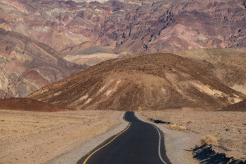 A highway in a barren desert landscape pointed towards a high wall of steep, colorful mountains