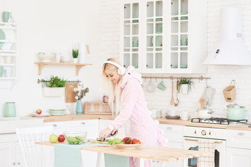Young Woman in cozy pajamas and headphones cooking in the kitchen and listening fun music. Healthy Food - Vegetable Salad. Diet. Dieting Concept. Healthy Lifestyle. Cooking At Home. 