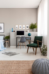 Grey pouf on natural linen carpet in fashionable teenager's room with retro armchair and industrial desk with computer