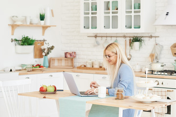 Freelance concept. Beautiful business woman in casual clothes and glasses is examining documents and smiling while working with a laptop in kitchen. Working at home