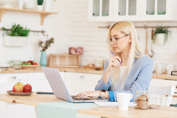 Freelance concept. Beautiful business woman in casual clothes and glasses is examining documents and smiling while working with a laptop in kitchen. Working at home