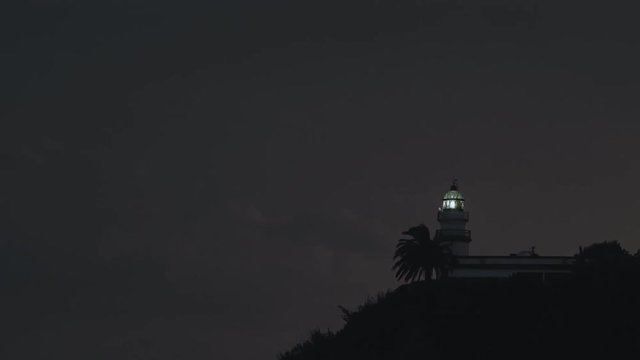 The sea lighthouse in Calella at night spins and gives rays of light against the background of lightning and thunderstorms.