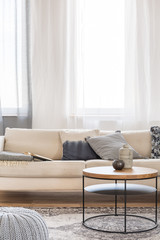 Round coffee table in front of beige sofa with pillows in bright living room interior