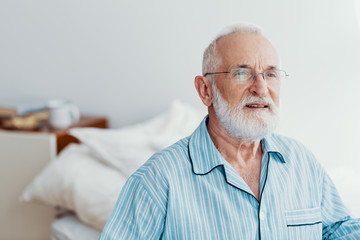 Old sick man with grey beard and hair wearing blue pajamas and sitting on bed at home
