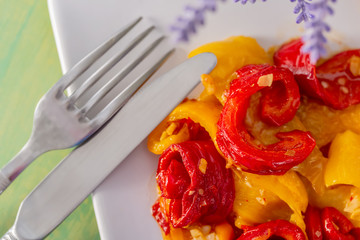 Cooked red yellow fried sweet peppers on a white plate. Fork and knife. Vegetarian food. Soft focus. Closeup. Top view.
