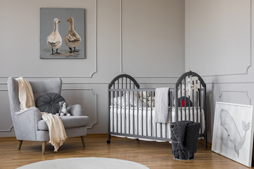 Comfortable grey armchair with round pillow and white blanket next to wooden crib with pillows and...