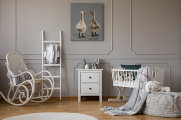 White wooden rocking chair with pillow next to scandinavian ladder, chest of drawers and cradle,...