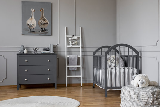 Stylish grey baby room interior with wooden furniture, white scandinavian ladder and teddy bear on pouf, real photo with copy space on the empty wall