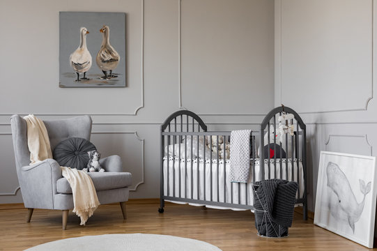 Comfortable grey armchair with round pillow and white blanket next to wooden crib with pillows and toys, copy space on empty wall