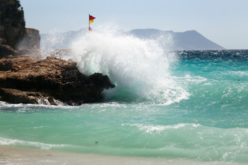 Waves coming from the sea are blasting on the rocks. Water color is turquoise and a red yellow flag is on the rock. Windy and stormy weather at Mediterranean Sea. Buyukcakil Beach, Antalya Turkey