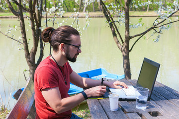 young modern man freelancer designer working outdoor by lake on laptop and tablet sunny spring summer day