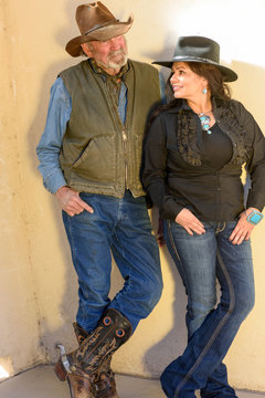 Old Cowboy with beard and pretty cowgirl with long, dark hair look at each other against a wall.
