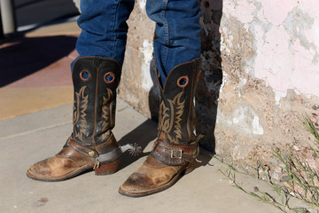 Old cowboy boots are weathered and brown with stitched design on the side with spurs worn by an old cowboy in jeans.
