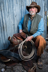 Old cowboy with gray beard wears leather hat, leather chaps, and holds a lasso and is sitting with a leather saddle.