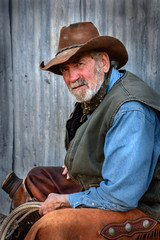 Old cowboy with gray beard wears leather hat, leather chaps and stares at camera. 