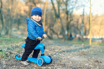 Cute adorable caucasian toddler boy in blue jacket having fun riding three-wheeled balance run bike scooter in city park or forest. Children outdoor sport activities