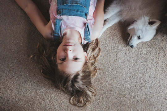 Overhead View Of Young Girl Lying On Carpet Floor With White Dog